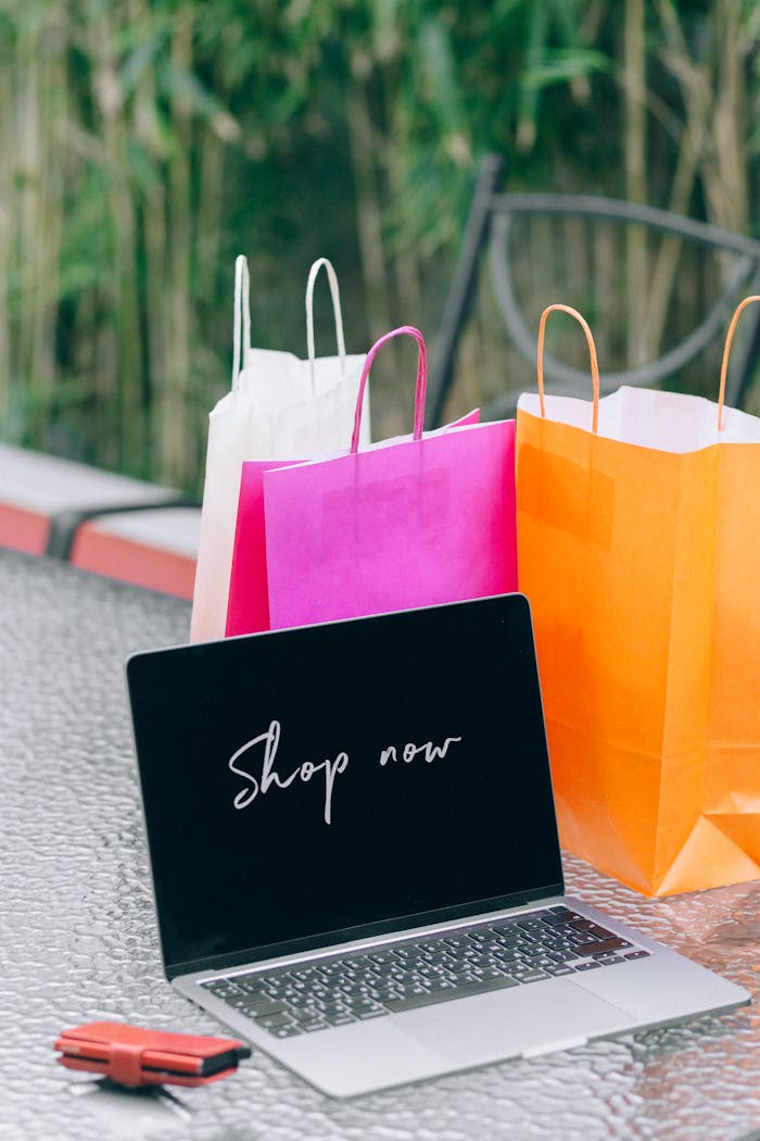 Laptop displaying Shop now with vibrant shopping bags on glass table outdoors.